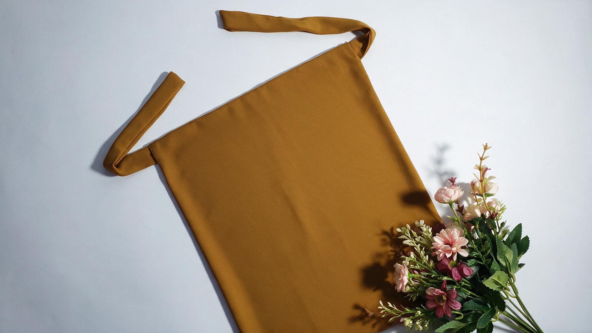 A mustard-coloured niqab laid flat and styled with a small bouquet of flowers, photographed the way a fashion product listing would present it.